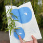 A photograph of Jo’s hand holding open the first spread of the book, their pinky finger gently pinning a single willow branch to the paper. The live branch emulates the illustration below, a line drawing of a similar willow tendril with a small seed looking up to a gibbous moon in a night sky. 