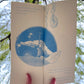 Hanson holds open the spread of the seedlings awakening from the soil. They have taken the photo of this zine from below, pointing up into the sky so that the blurry canopy of a large willow tree wraps around the open spread, filling the background of the photo. 