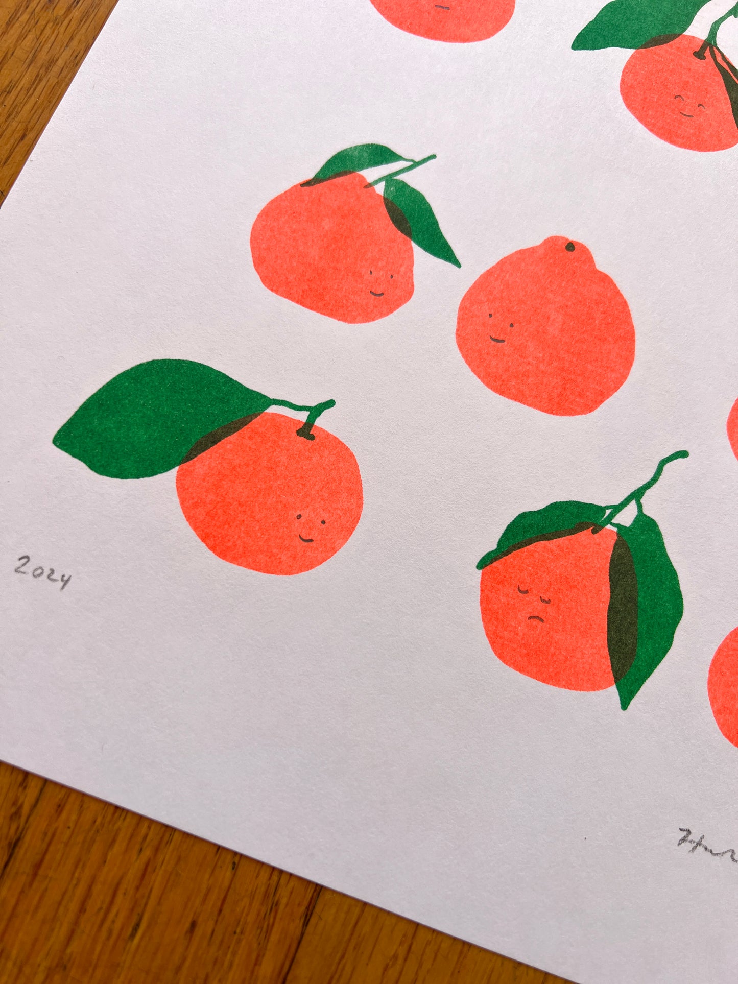 A close up photo of the lower left corner of the print, showing the hand penciled year "2024". Five of the printed satsumas and be seen, as well as the Risograph texture and color overlay of the green leaves printing atop the fluorescent orange peels. 