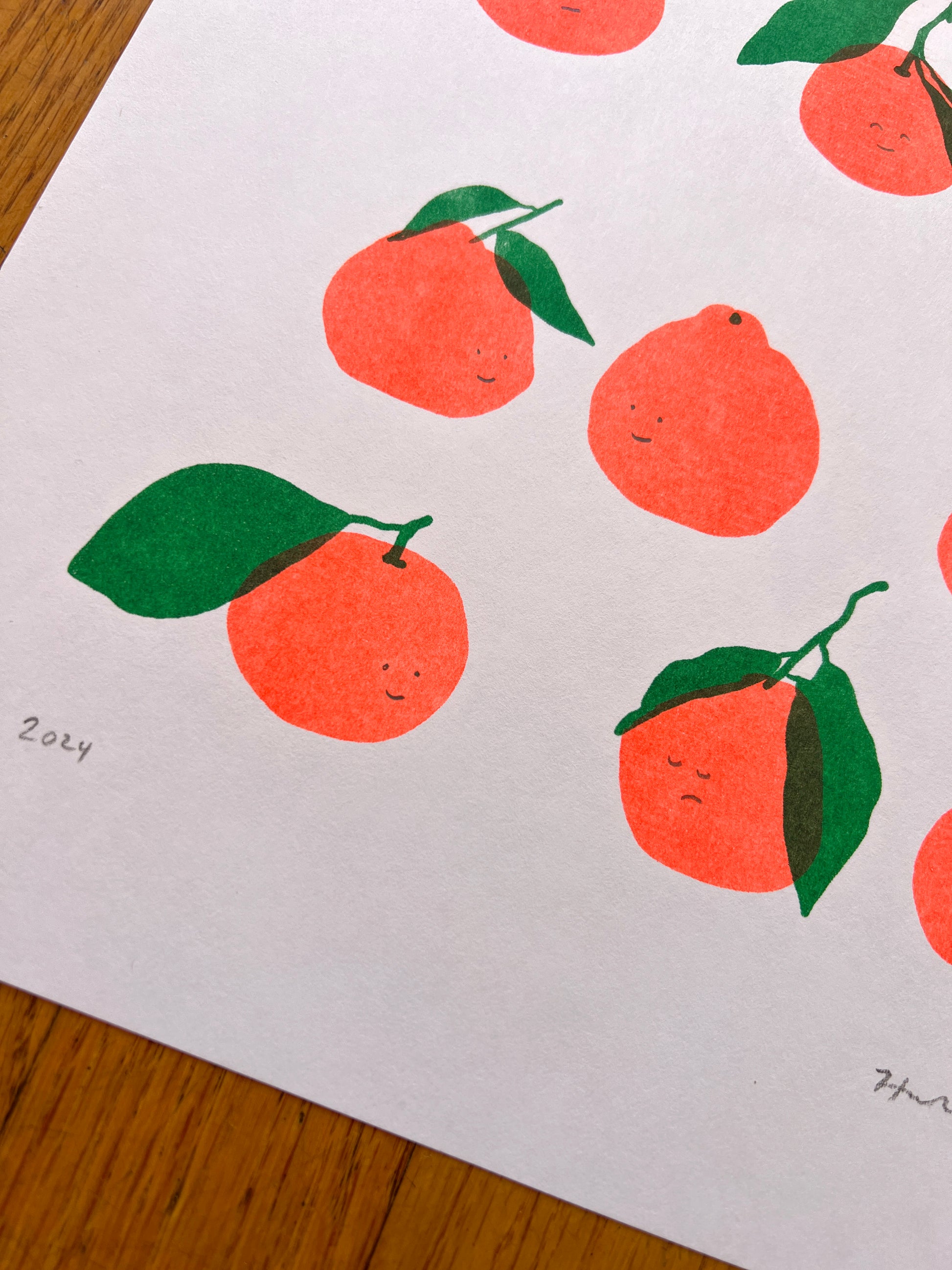 A close up photo of the lower left corner of the print, showing the hand penciled year "2024". Five of the printed satsumas and be seen, as well as the Risograph texture and color overlay of the green leaves printing atop the fluorescent orange peels. 