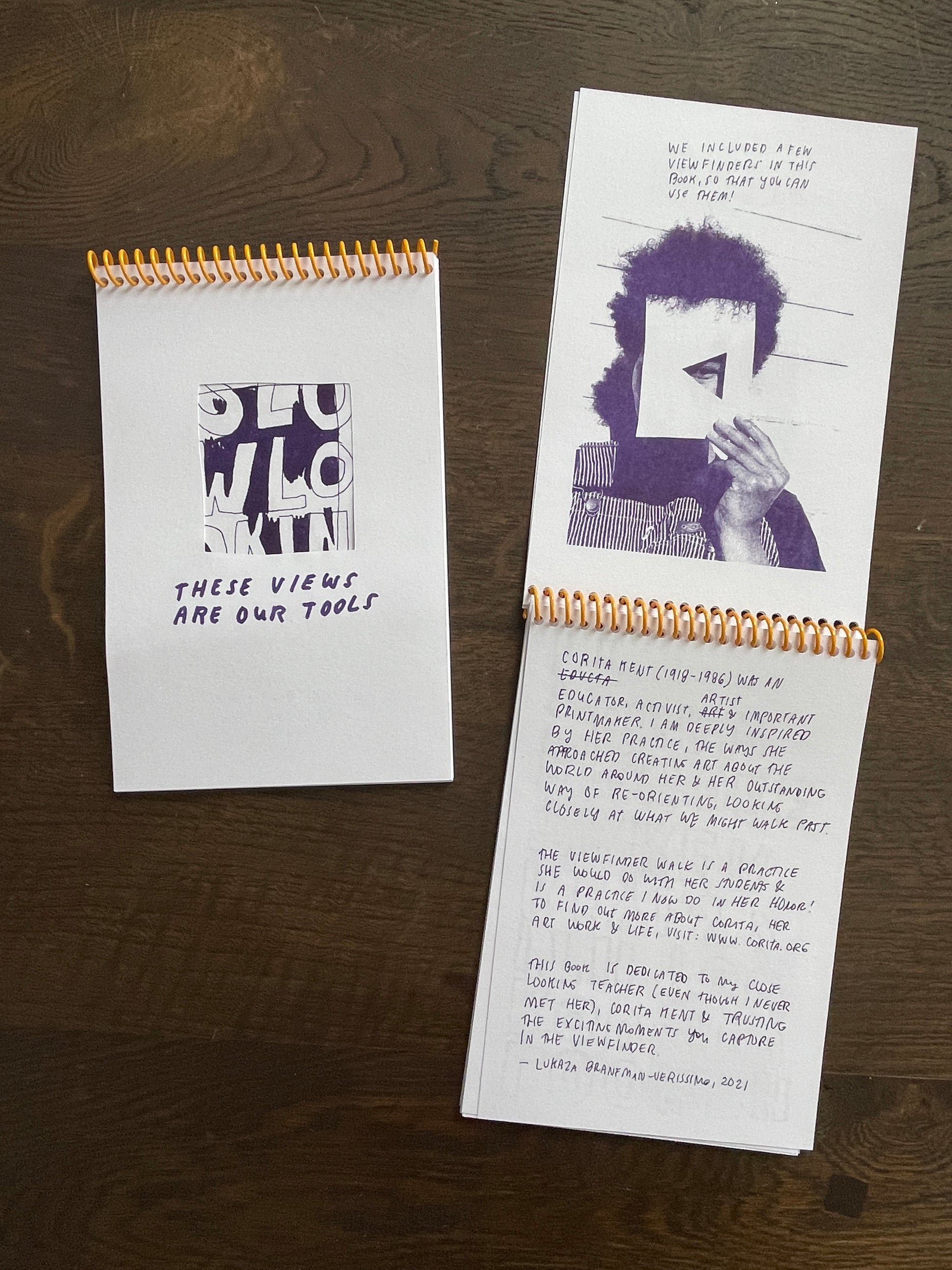 Two copies of the same zine rest on a dark wooden table top. The right is closed showing on the cover. The left zine is opened to a vertical spread Risograph printed on white paper in purple ink. The top page has an image of a person holding a viewfinder to their face revealing just one eye. The bottom spread includes handwritten information about the artist Corita Kent. 