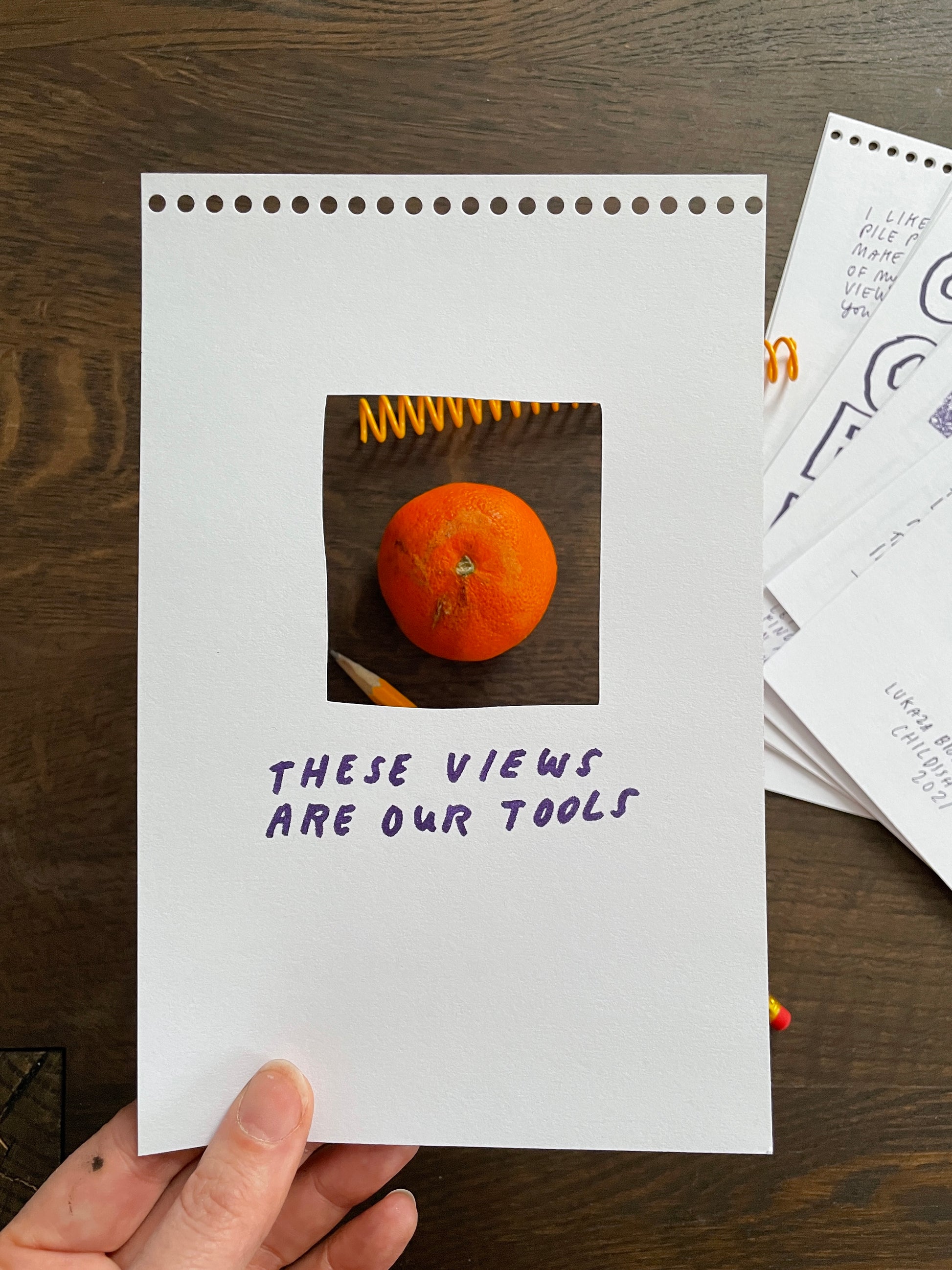 A photo of the book Slow Looking set on a table unbound from it’s coil. On the right The interior pages are slightly fanned out on the dark wooden surface. Towards the left, a white hand holds the cover sheet a few inches above the table. The center of the cover has a die-cut rectangle which frames a bright orange satsuma, the books loose yellow coil, and a number 2 pencil, on the table below. 